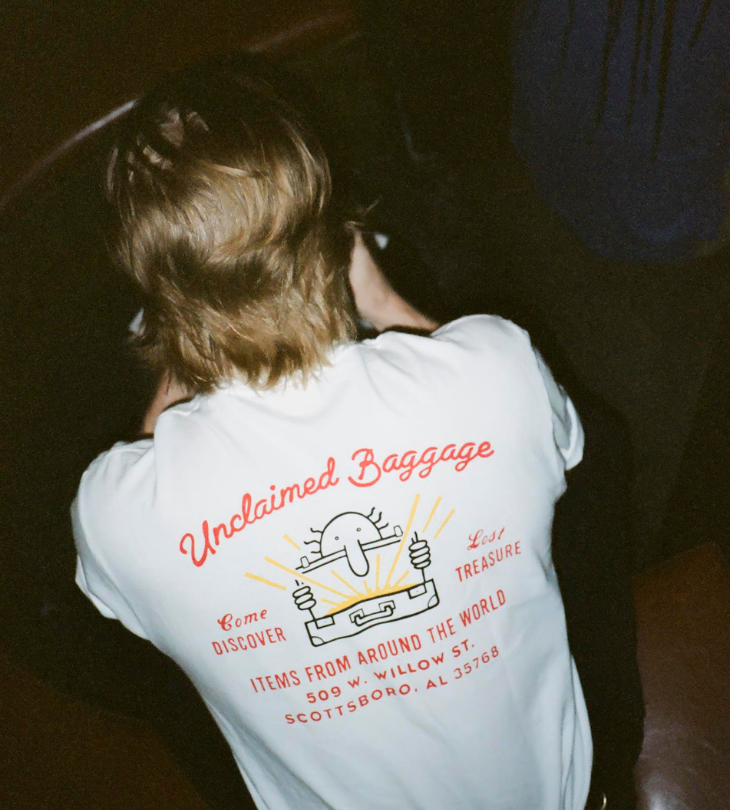 a person sitting down with their back facing wearing a Unclaimed Baggage white t-shirt